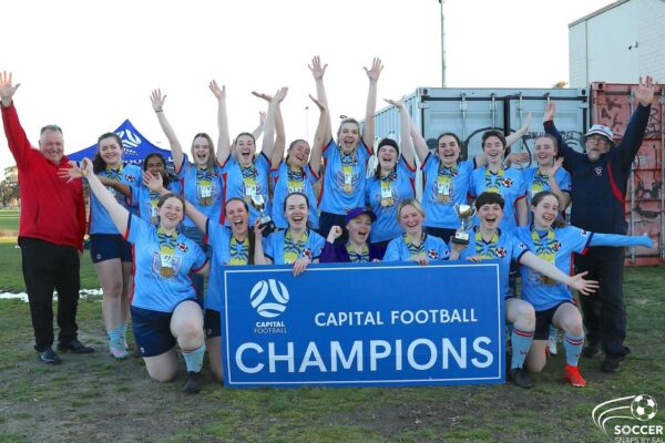 A young woman's soccer team and two coaches smiling behind the billboard, "Capital Football Champions"