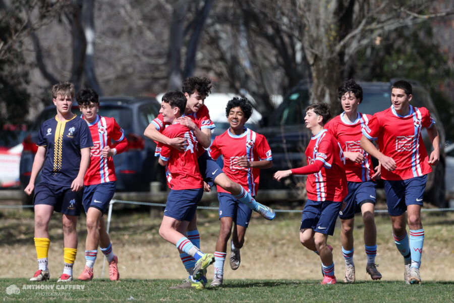A team celebrates after a goal, with two boys hugging in the foreground while four teammates trail behind, smiling and running toward them.